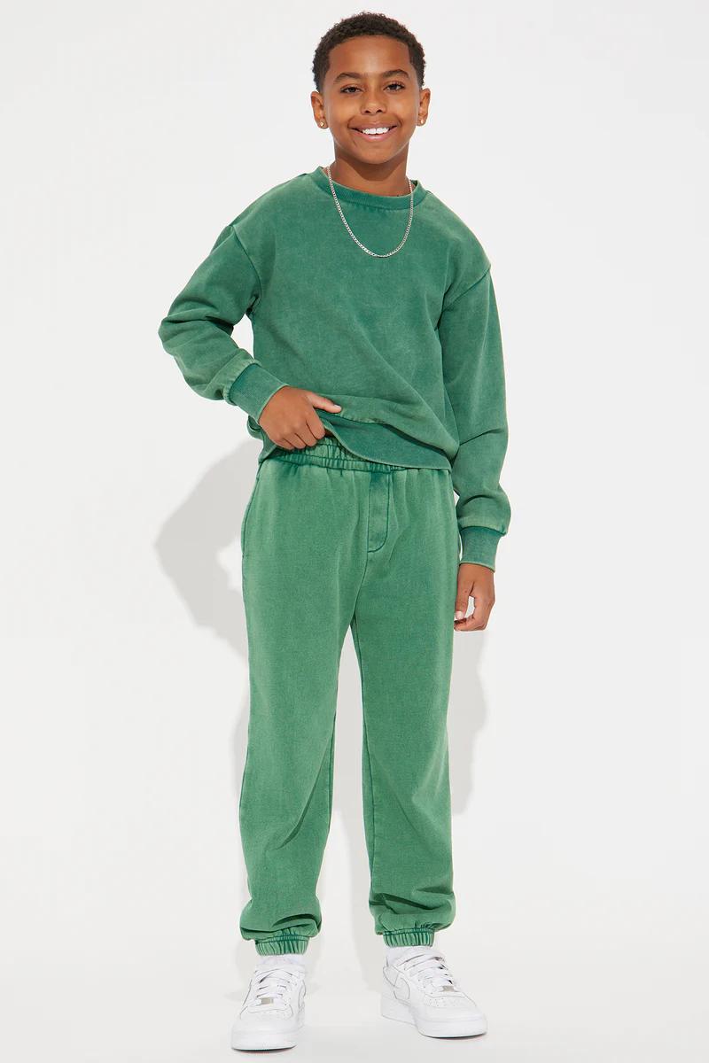 Smiling boy wearing a matching kids green sweatshirt and sweatpants jogger set with white sneakers.