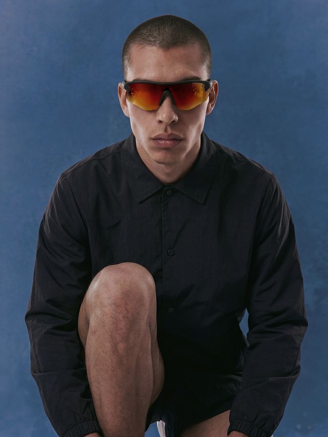 Man in black jacket, shorts, and orange reflective performance sunglasses, crouching against a blue background. Showcases new athletic eyewear collection.