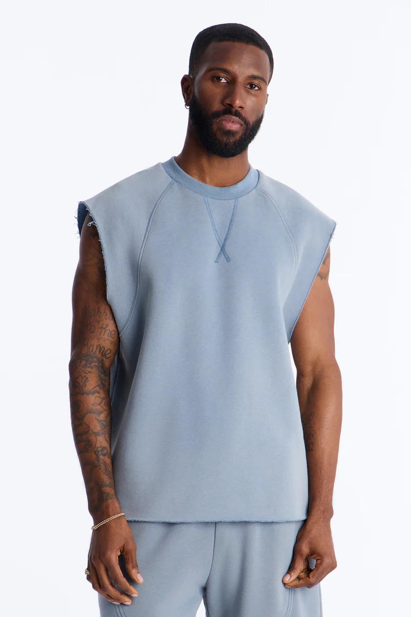 Man models dusty blue sleeveless crewneck sweatshirt and sweatpants, showcasing comfortable men's raw edge athleisure.
