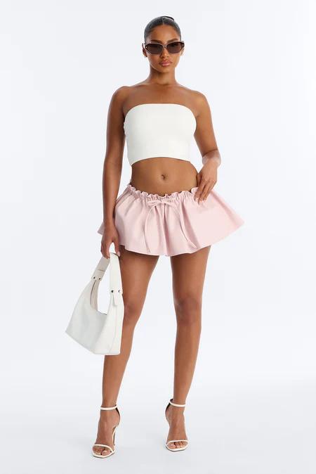 Modelo con top blanco sin tirantes, minifalda rosa con volantes, tacones blancos, bolso de hombro blanco y gafas de sol.