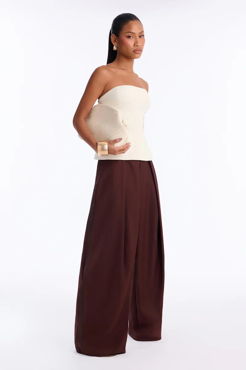 Woman wearing a cream strapless top and brown wide-leg trousers holding a clutch.