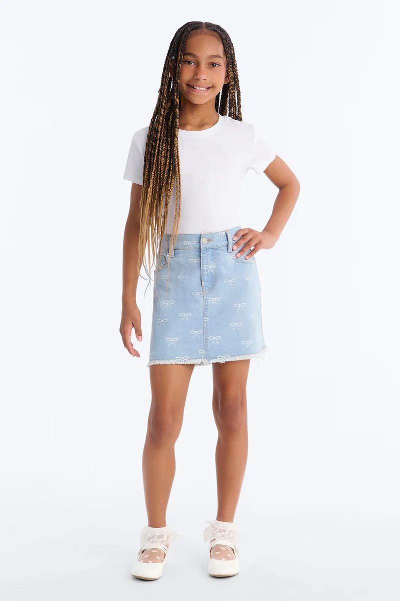 Niña sonriente, camiseta blanca, minifalda vaquera azul con lazos y bajo deshilachado. Zapatos mary jane blancos.