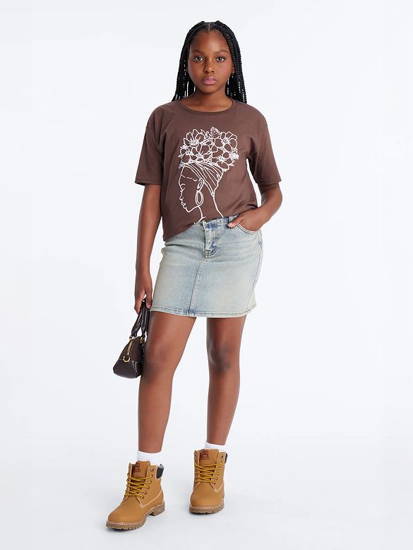 Young girl in a brown graphic tee with a white floral design, light wash denim skirt, tan boots, and dark bag. Shop kids' fashion.