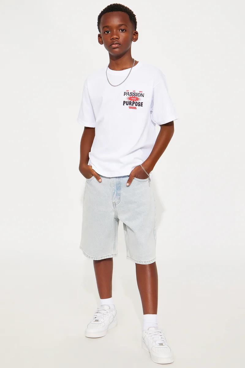 Young boy models a white "Passion and Purpose" graphic tee, light denim shorts, and white sneakers. Stylish kids' casual outfit.