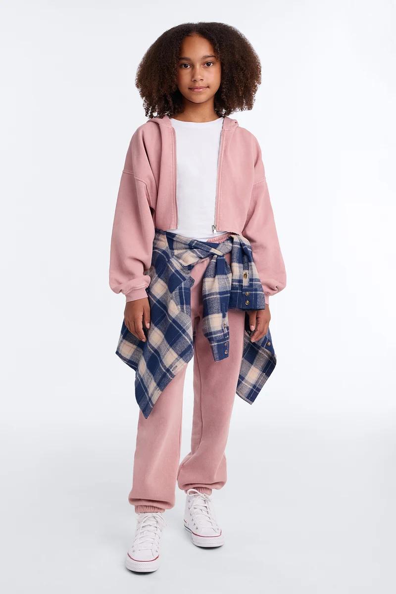 Niña con sudadera corta rosa, pantalones deportivos a juego, camisa de franela a cuadros y tenis blancos.