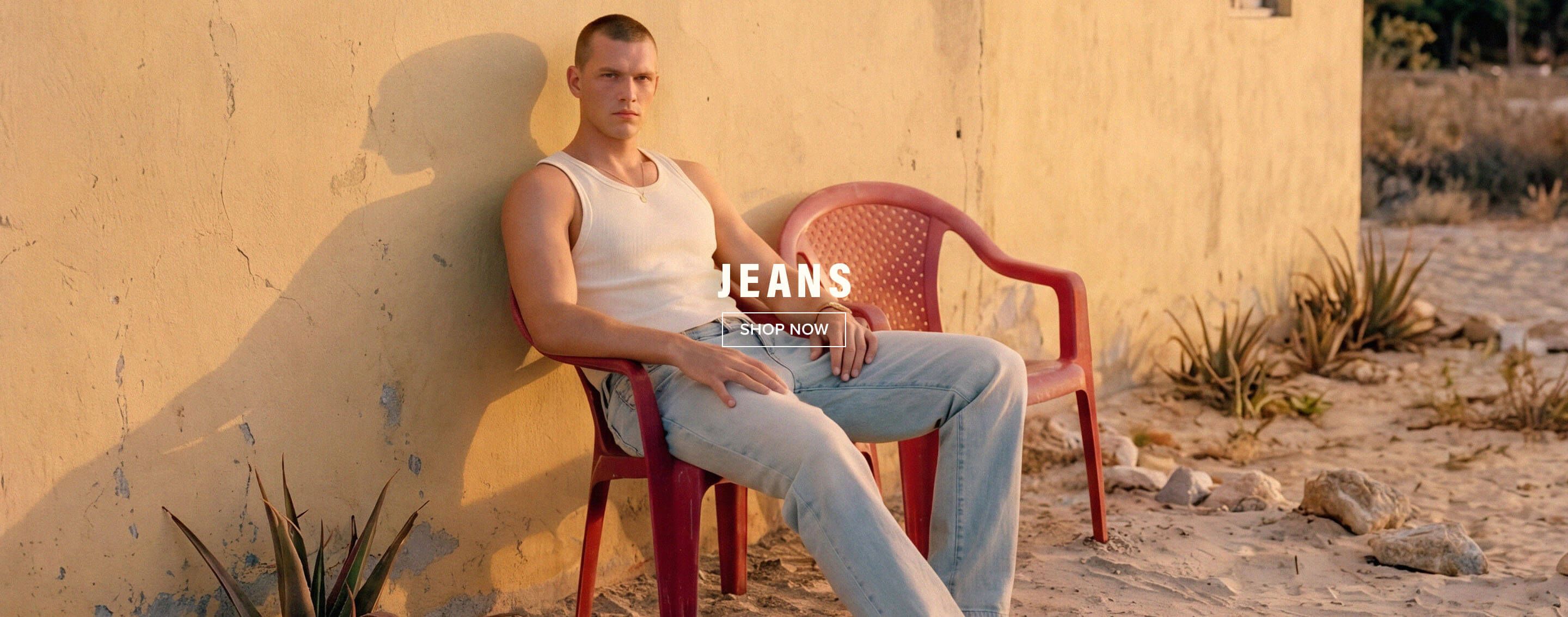 Man in a white tank top and light blue jeans seated on a red chair against a yellow wall. Shop the new men's jeans collection now.