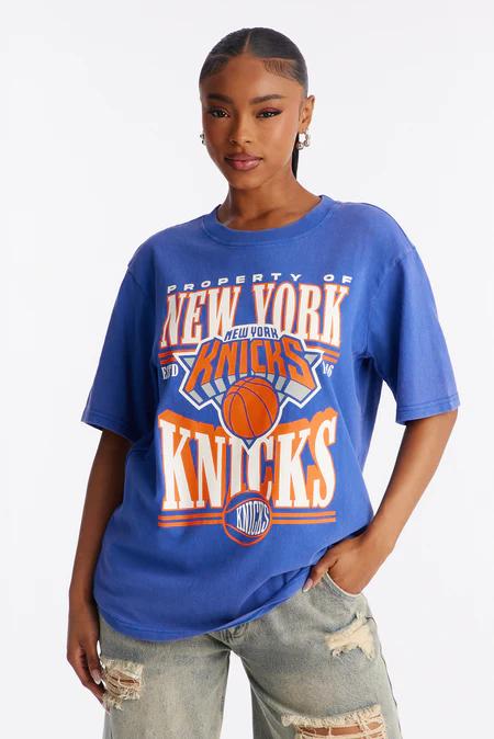 Woman in a white one-shoulder crop top with yellow trim and side ruching, featuring the Lakers logo. She wears baggy light wash jeans and silver bangles.
