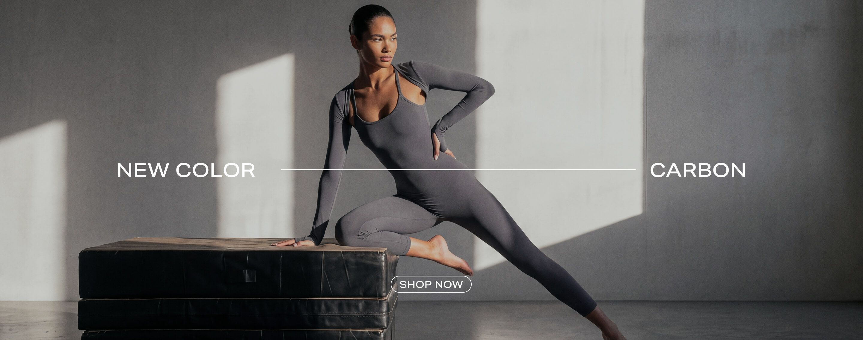 A woman poses in a sleek carbon grey long-sleeve activewear bodysuit on a black block, showcasing the brand's new carbon colorway.