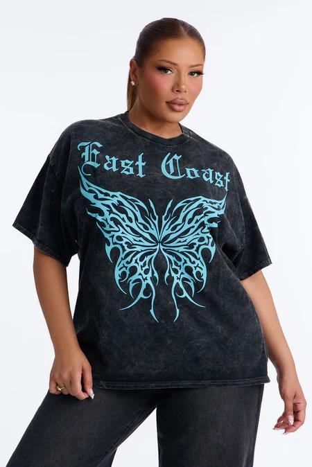 Woman models an oversized washed black t-shirt with bold blue 'East Coast' text and a tribal butterfly graphic. She wears dark jeans.