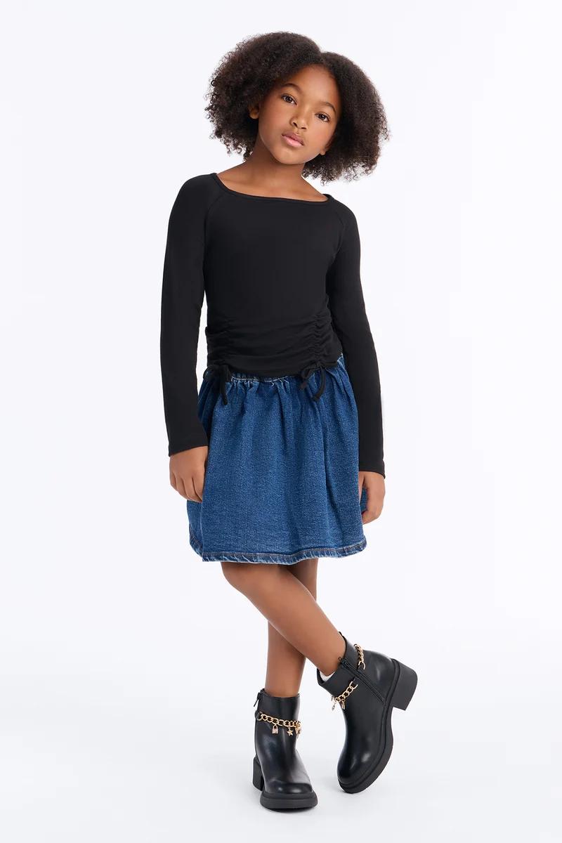 Girl wearing a long-sleeve black ruched top, blue denim skirt, and black ankle boots with gold chains.