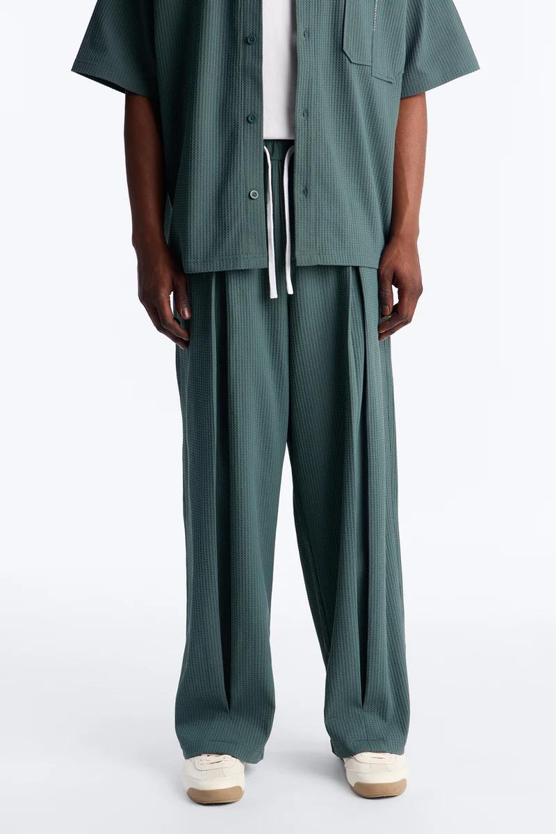 Men's dark teal waffle knit short-sleeve shirt and pleated drawstring pants with cream sneakers. Highlighting new comfortable casual wear.