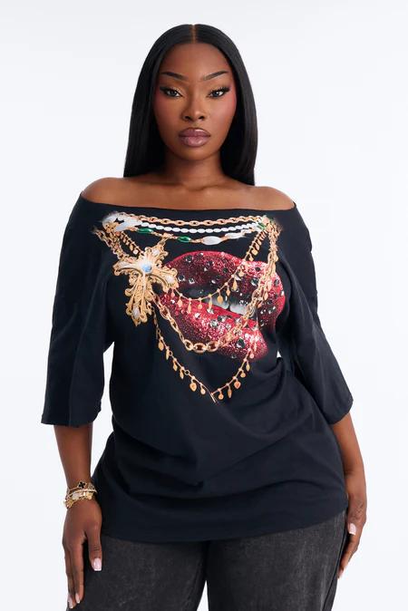 Mujer con top negro de hombros descubiertos y gráfico de labios rojos brillantes, adornado con cadenas doradas, perlas y una cruz enjoyada.