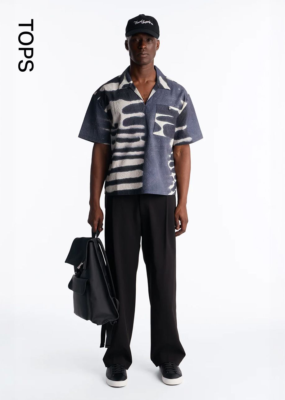 Man wearing a black cap, blue and white patterned casual short-sleeve shirt, black trousers, holding a backpack. Shop men's stylish tops.