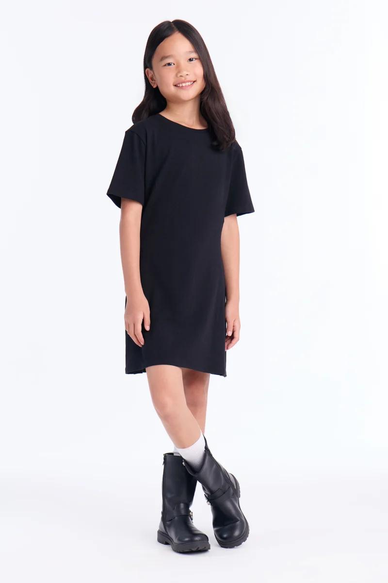 Niña sonriente con vestido tipo camiseta negro casual, calcetines y botas negras.