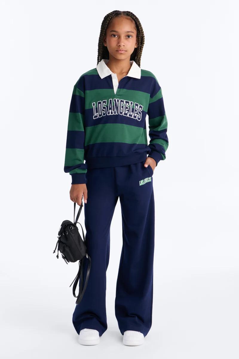 Girl in green and navy striped Los Angeles rugby shirt and matching wide-leg sweatpants holding a black backpack.