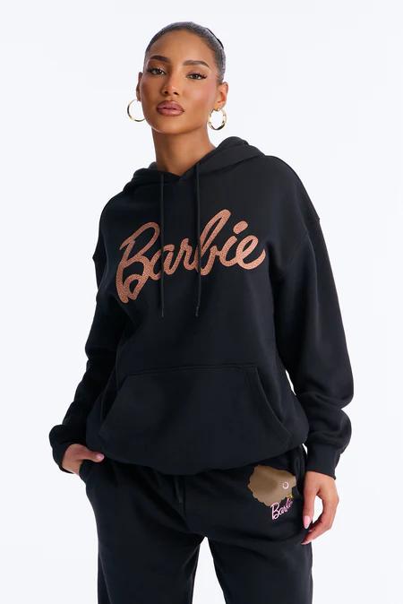 Woman in a black hoodie with "Barbie" in glittery rose gold script. Matching black sweatpants feature a subtle Barbie head graphic.