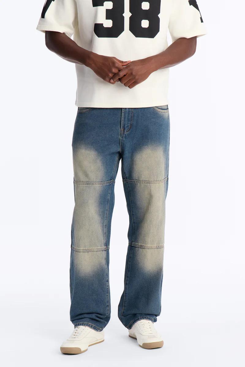 Persona vistiendo camiseta gráfica crema con "38" y vaqueros azules anchos desgastados con paneles, promocionando nueva moda masculina.