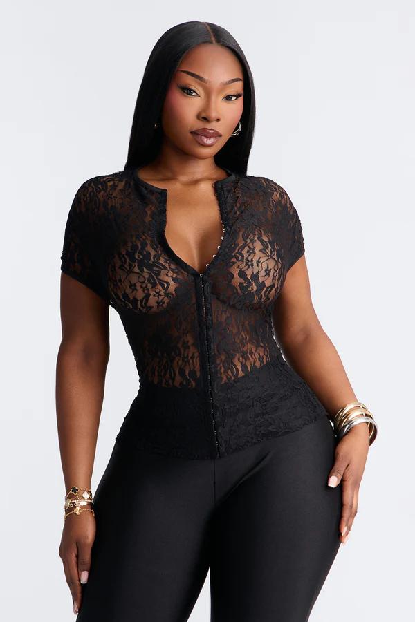 Woman in a black sheer lace short-sleeved top with a hook-and-eye front closure, black pants, and gold bracelets. White background.