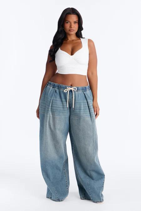 Mujer con cabello oscuro largo, top corto blanco fruncido y pantalones anchos de mezclilla azul claro con cordón, sobre fondo blanco.