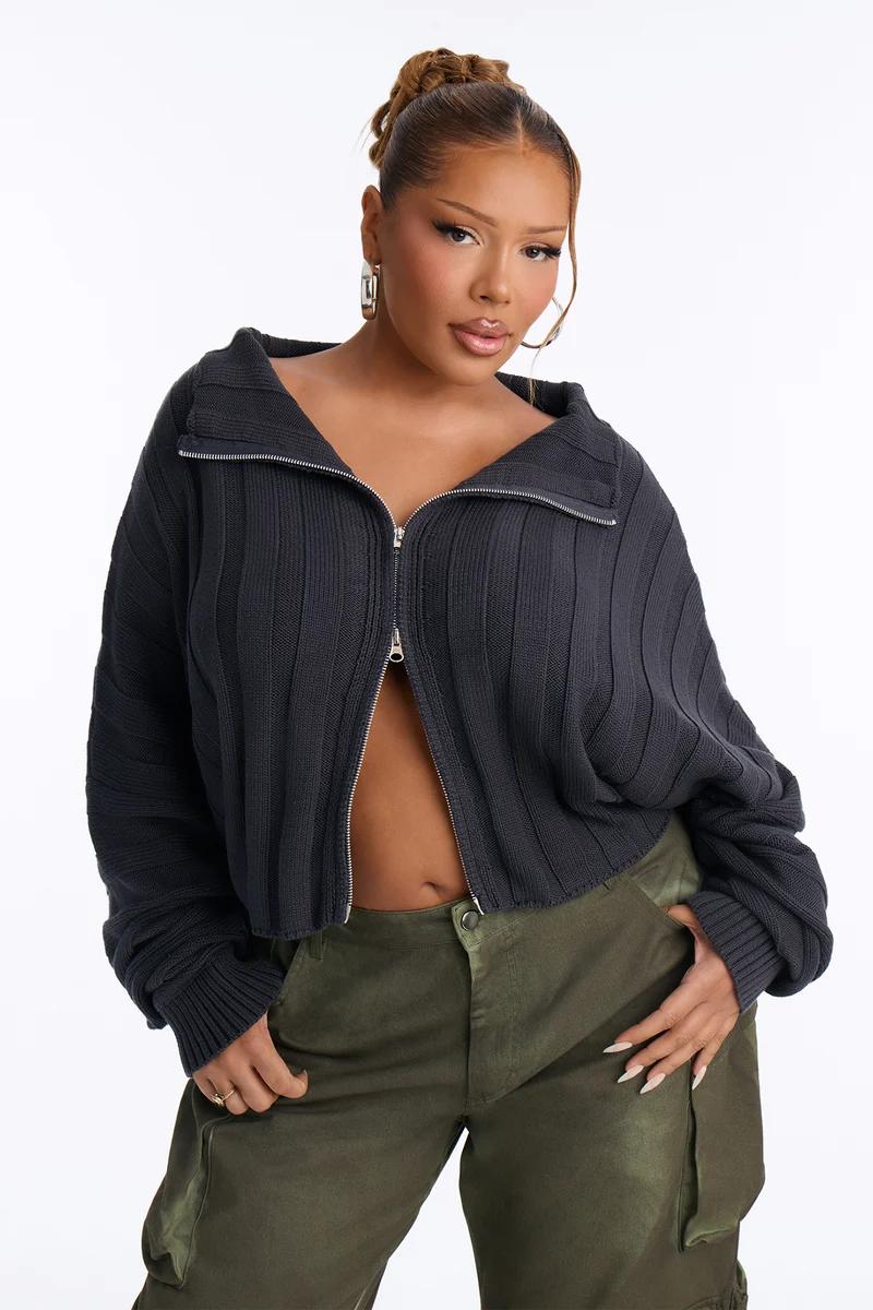 Plus size woman in a cropped charcoal grey ribbed zip-up sweater and olive green cargo pants.