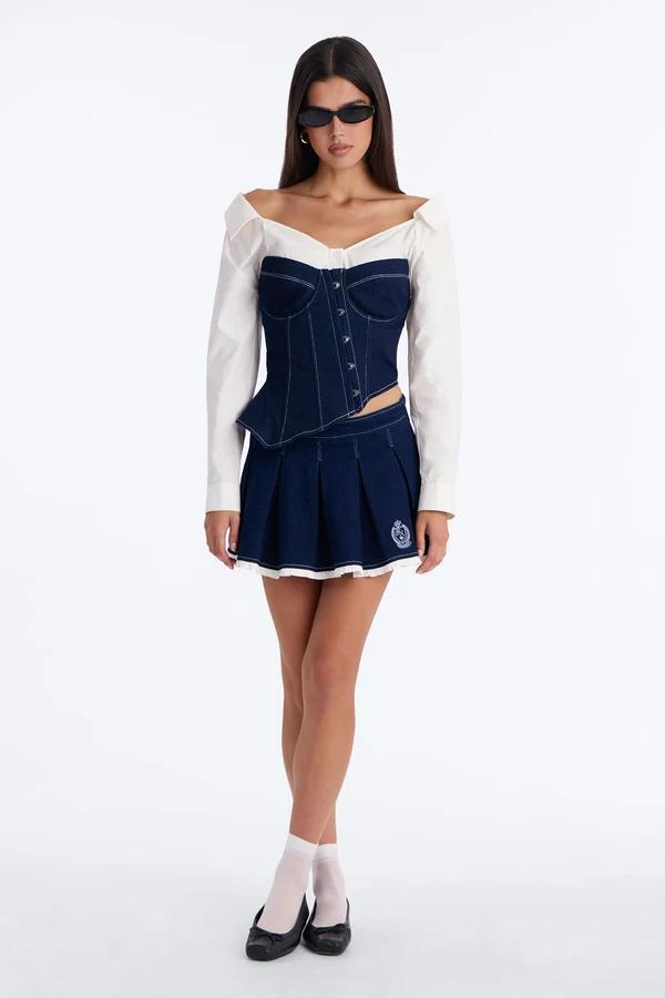 Model wearing a dark denim corset top over a white off-shoulder shirt and matching pleated mini skirt, promoting a preppy fashion collection.