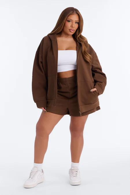 Full-body shot of a woman in an oversized brown zip-up hoodie, matching shorts, a white tube top, and white sneakers.