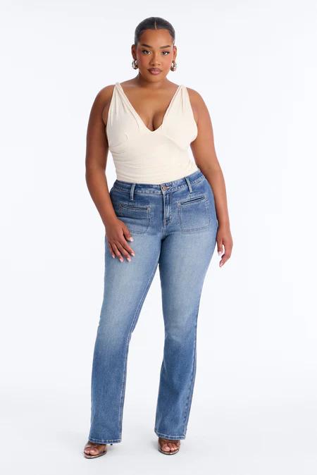 Una mujer con un top beige claro de cuello en V y jeans azules de lavado medio con bolsillos, de pie sobre fondo blanco.