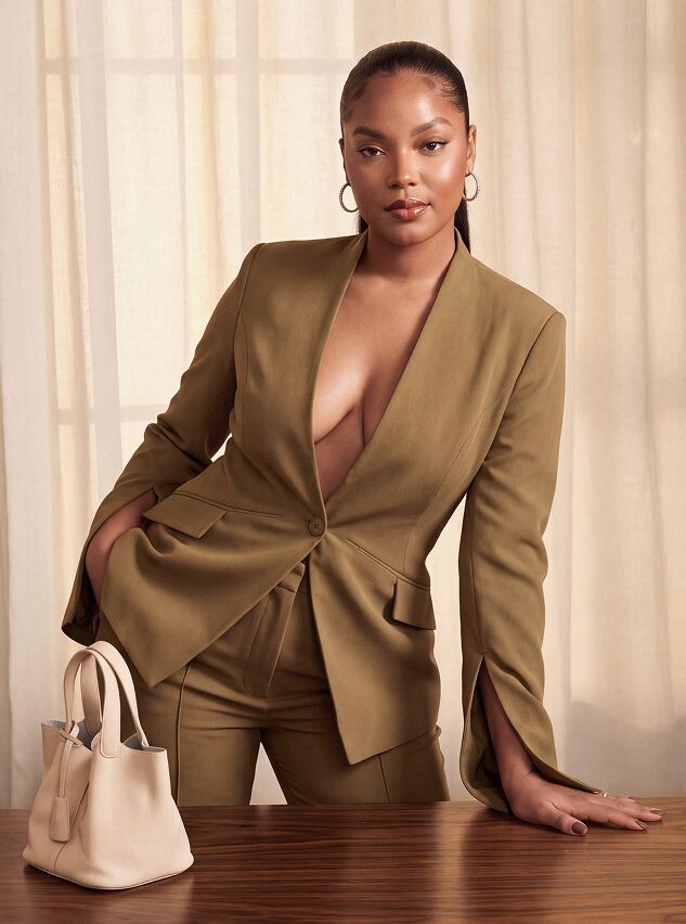 Mujer con elegante traje sastre verde oliva se apoya en mesa de madera junto a bolso de cuero crema, mostrando moda profesional moderna.