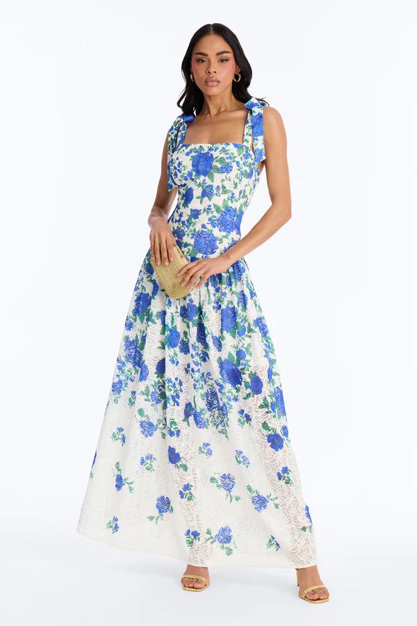 Woman in a square-neck, white maxi dress with blue floral print, bow straps, and a lace hem, holding a straw clutch.