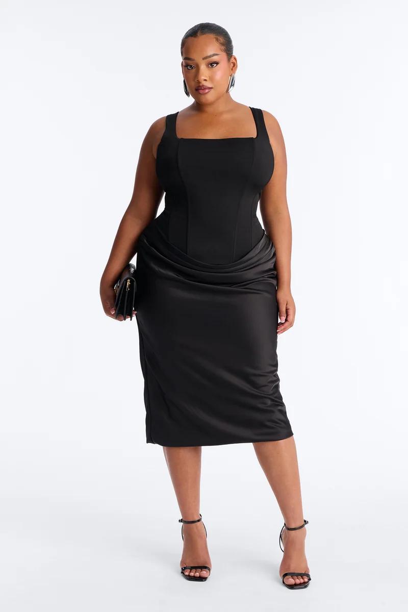 Woman wearing an elegant plus size black corset midi dress with draped skirt details, paired with black heels for a chic evening collection.