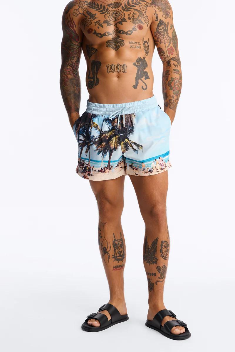 Man in light blue tropical beach print swim shorts with palm trees and black sandals. Ideal men's summer beachwear.