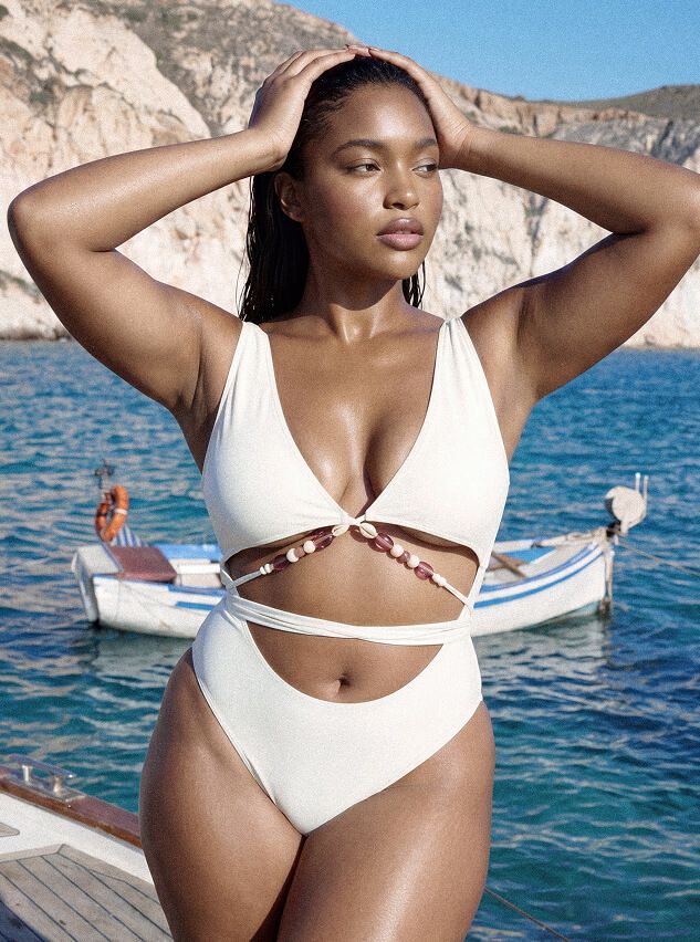 Modelo con traje de baño blanco de una pieza con recortes y cuentas, posando en mar turquesa y acantilados. Explora nueva moda de playa.