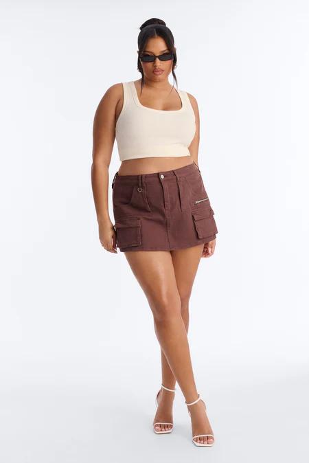 Woman in a cream square-neck crop top, dark brown cargo mini skirt with pockets, and white strappy heels. She wears narrow sunglasses.