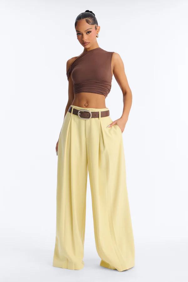 Modelo con top asimétrico marrón de un hombro, pantalones anchos plisados amarillo pálido y cinturón marrón con hebilla dorada.