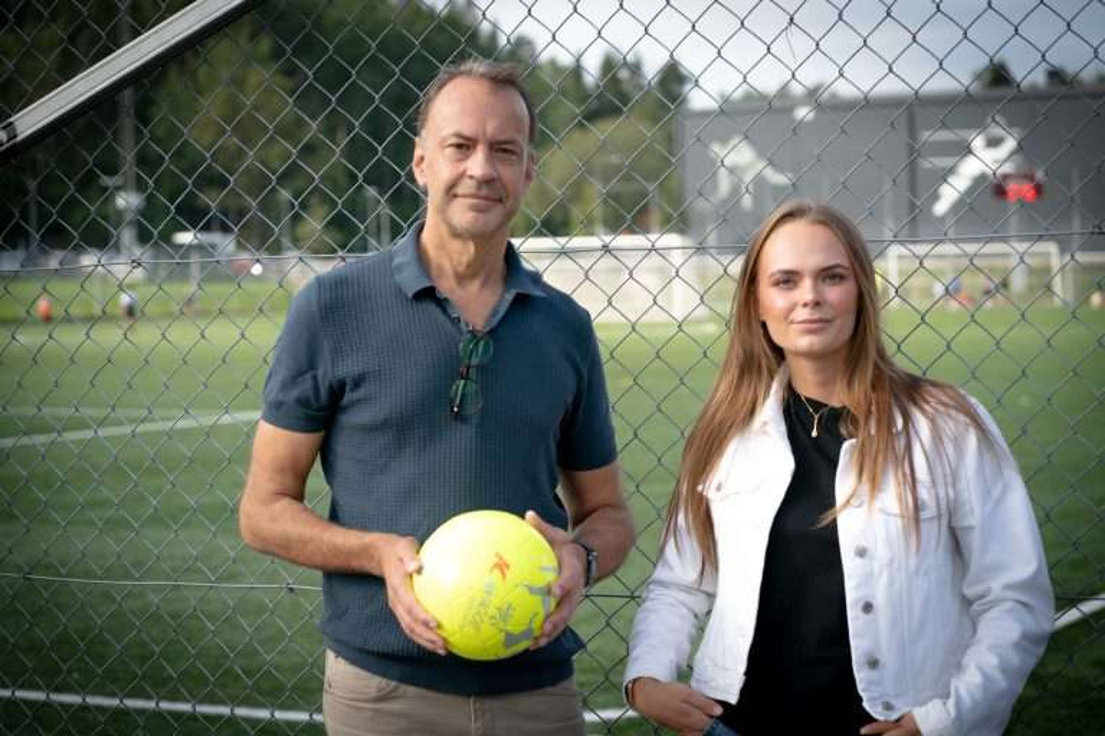 Två personer står vid ett stängsel intill en fotbollsplan. Den ena håller i en gul fotboll och i bakgrunden syns planen och mål