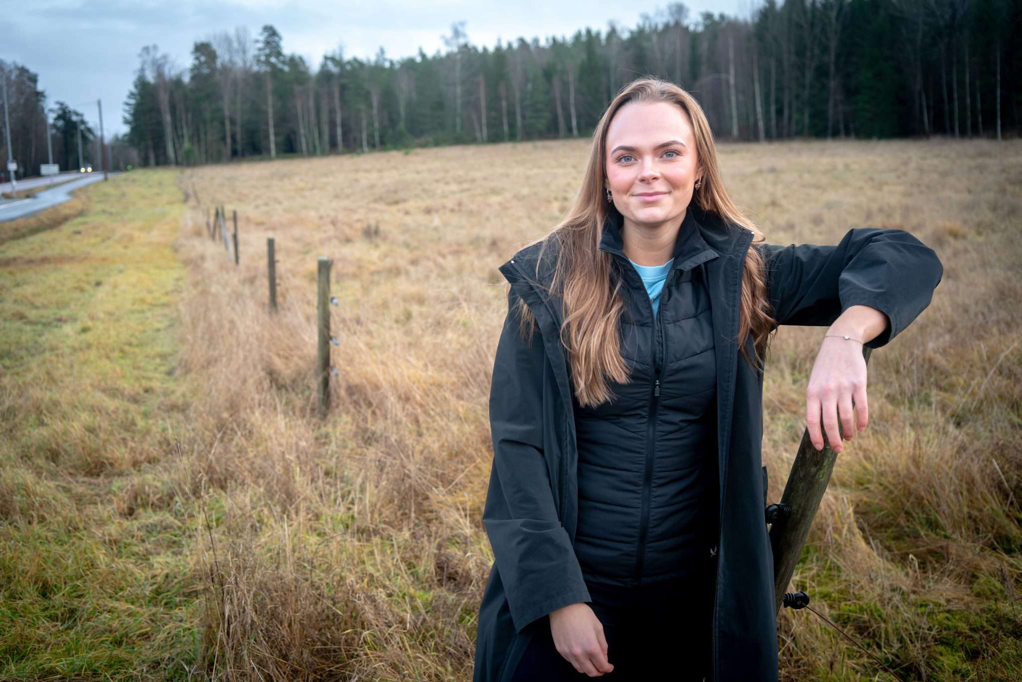 En kvinna står vid ett stängsel på en öppen äng i Källtorp där en ny fotbollsplan planeras att byggas. I bakgrunden syns skog och en väg