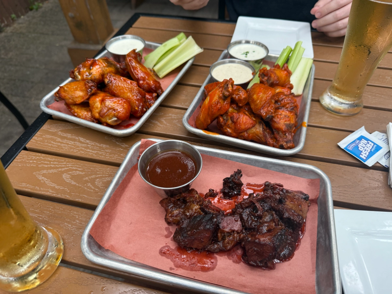 Two trays of saucy chicken wings with celery sticks and cups of creamy dip on an outdoor wooden table.