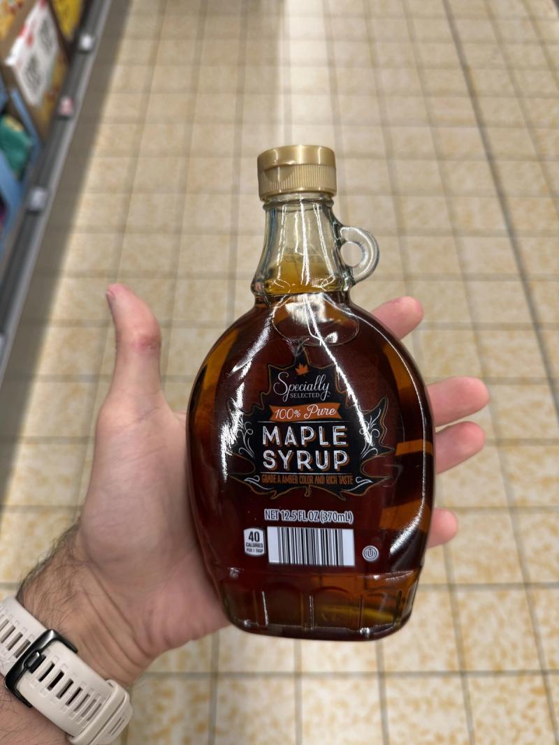 A hand holding a small glass jug of Specially Selected 100% Pure Maple Syrup in a grocery store aisle