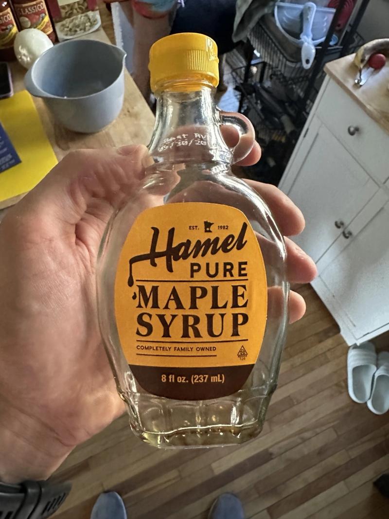 A hand holding a nearly empty 8oz bottle of Hamel pure maple syrup in a kitchen