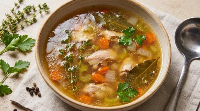 A jar of clear homemade chicken stock surrounded by fresh root vegetables.