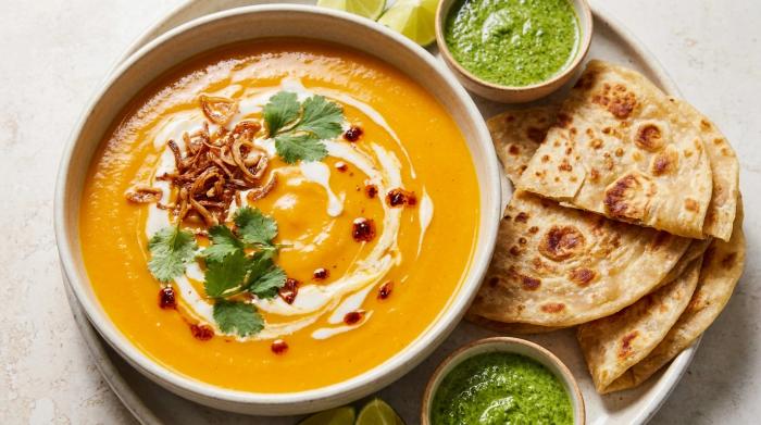 Close-up of vibrant orange Thai coconut pumpkin soup topped with a coconut milk swirl, fried shallots, and fresh herbs.