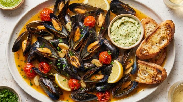 A wide overhead shot of steamed mussels in a white wine broth served with bread and lemon.