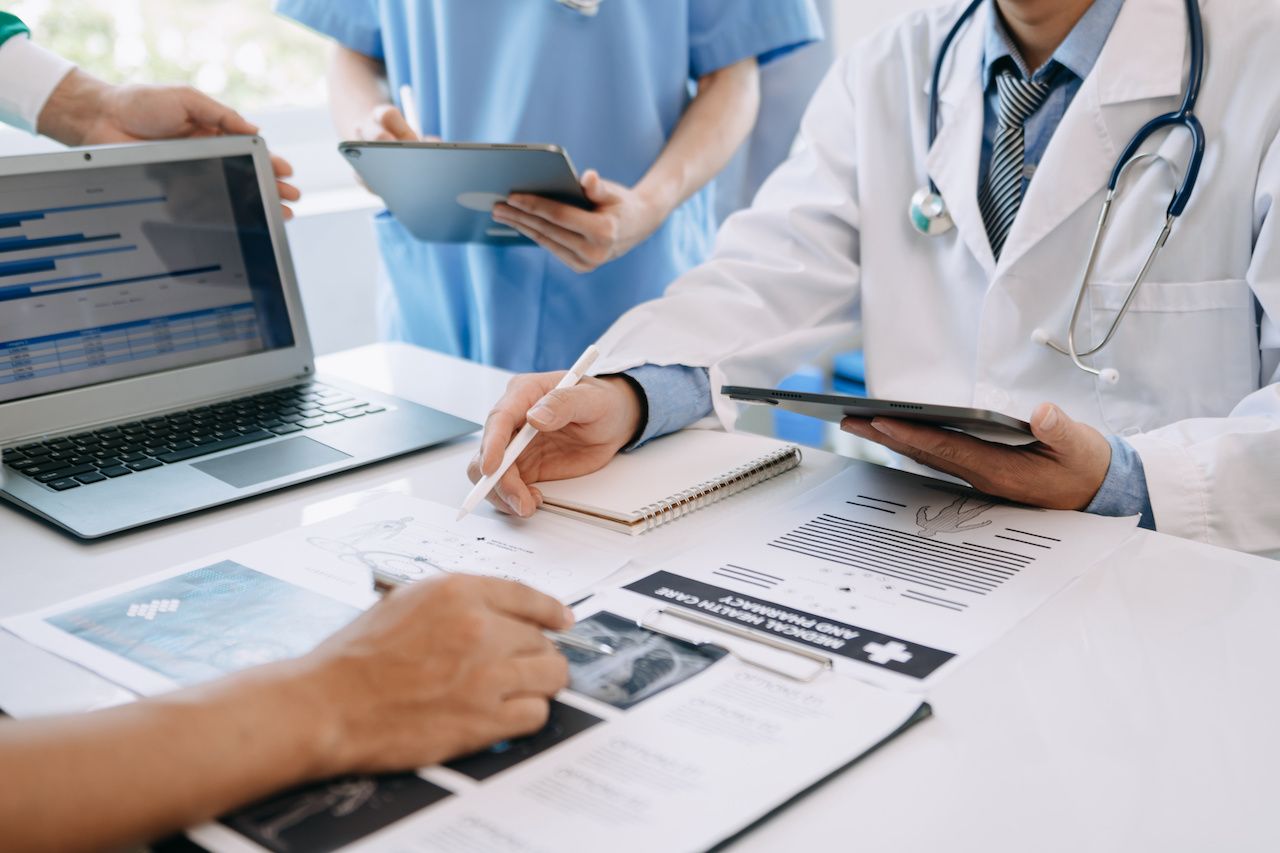 Medical team having a meeting with doctors in white lab coats and surgical scrubs seated at a table discussing a patients working online using computers in medical industry: © mrmohock - stock.adobe.com