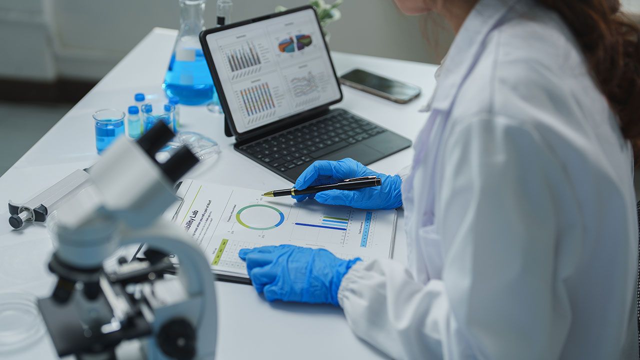Scientist wearing blue gloves pointing at charts and graphs on a paper document while analyzing medical research data using a tablet in a laboratory setting, with a microscope and beakers nearby | Image Credit: ©crizzystudio – stock.adobe.com