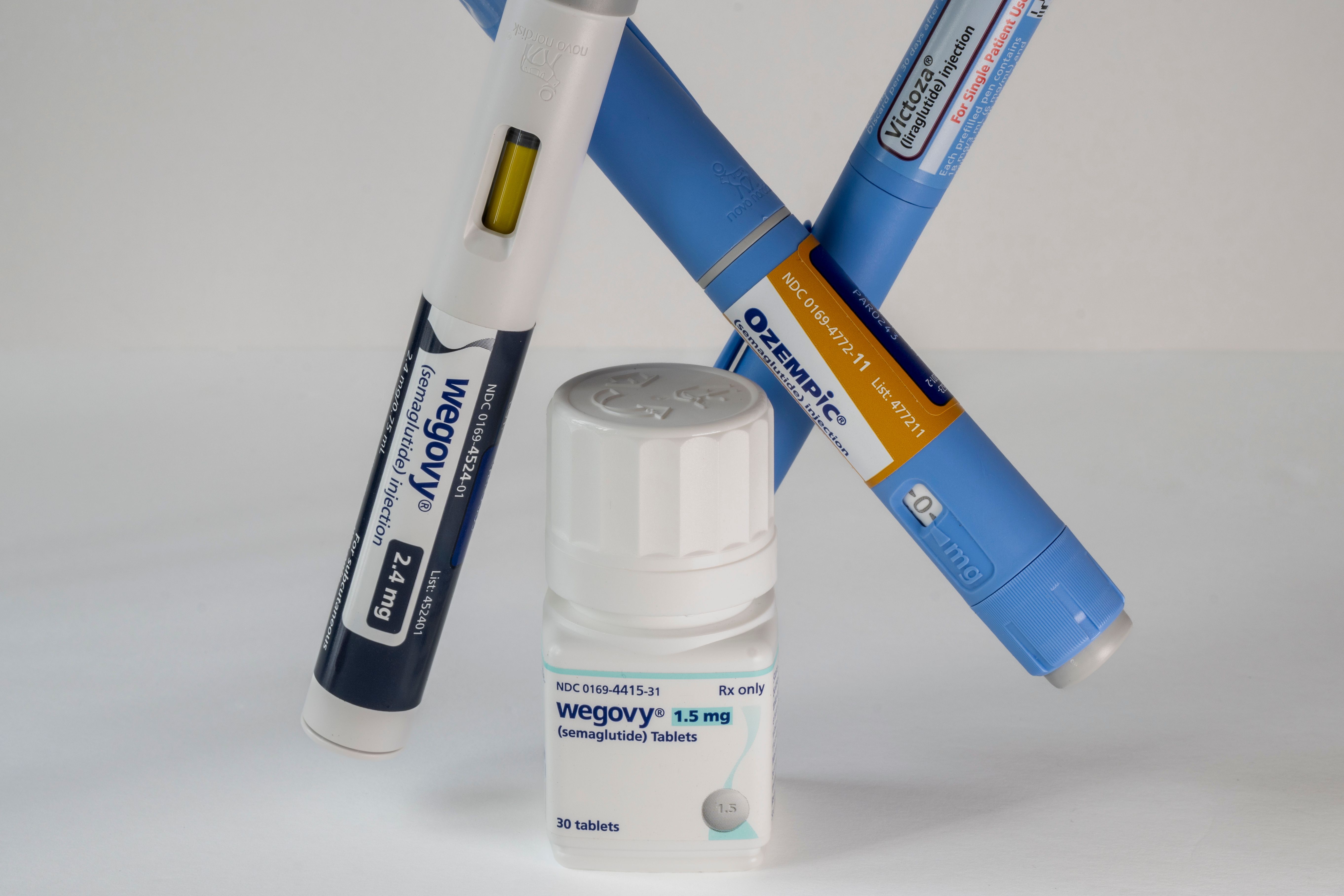Three injection pens labeled Wegovy, Ozempic, and Victoza crossed behind a bottle of Wegovy semaglutide 1.5 mg tablets on a white background. | Image Credit: © silukstockimages - stock.adobe.com