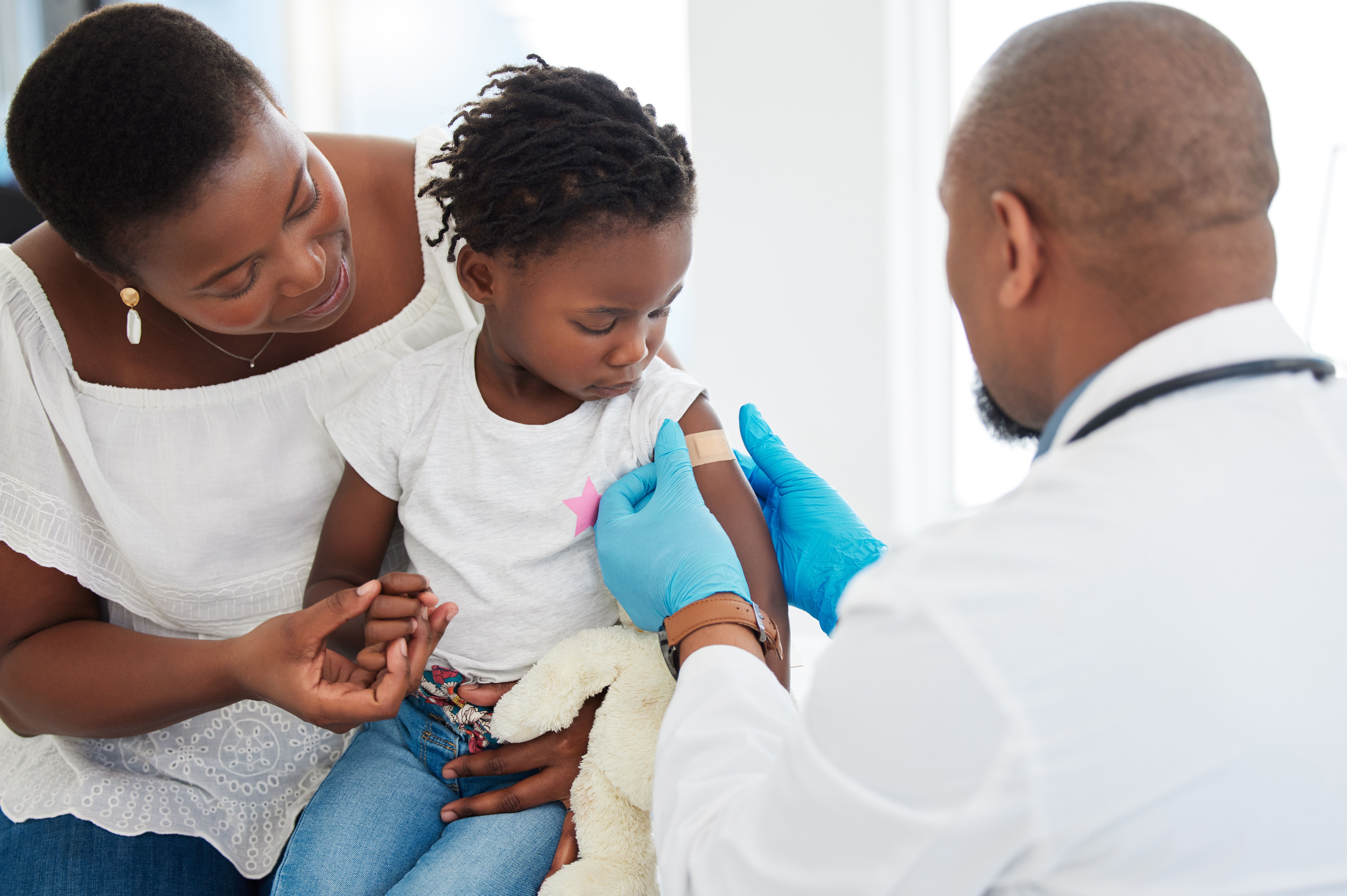 Photograph of a young girl with a bandage on her arm after receiving a vaccine, with her mother and a pediatrician present. | Image Credit: © Nina Lawrenson/peopleimages.com - stock.adobe.com