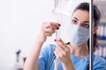 A woman in light blue scrubs and a surgical mask holding the drip chamber below a clear IV bag hanging from a metal pole, with a stethoscope around her neck and a blurred office background. | Image Credit: © Elnur - stock.adobe.com