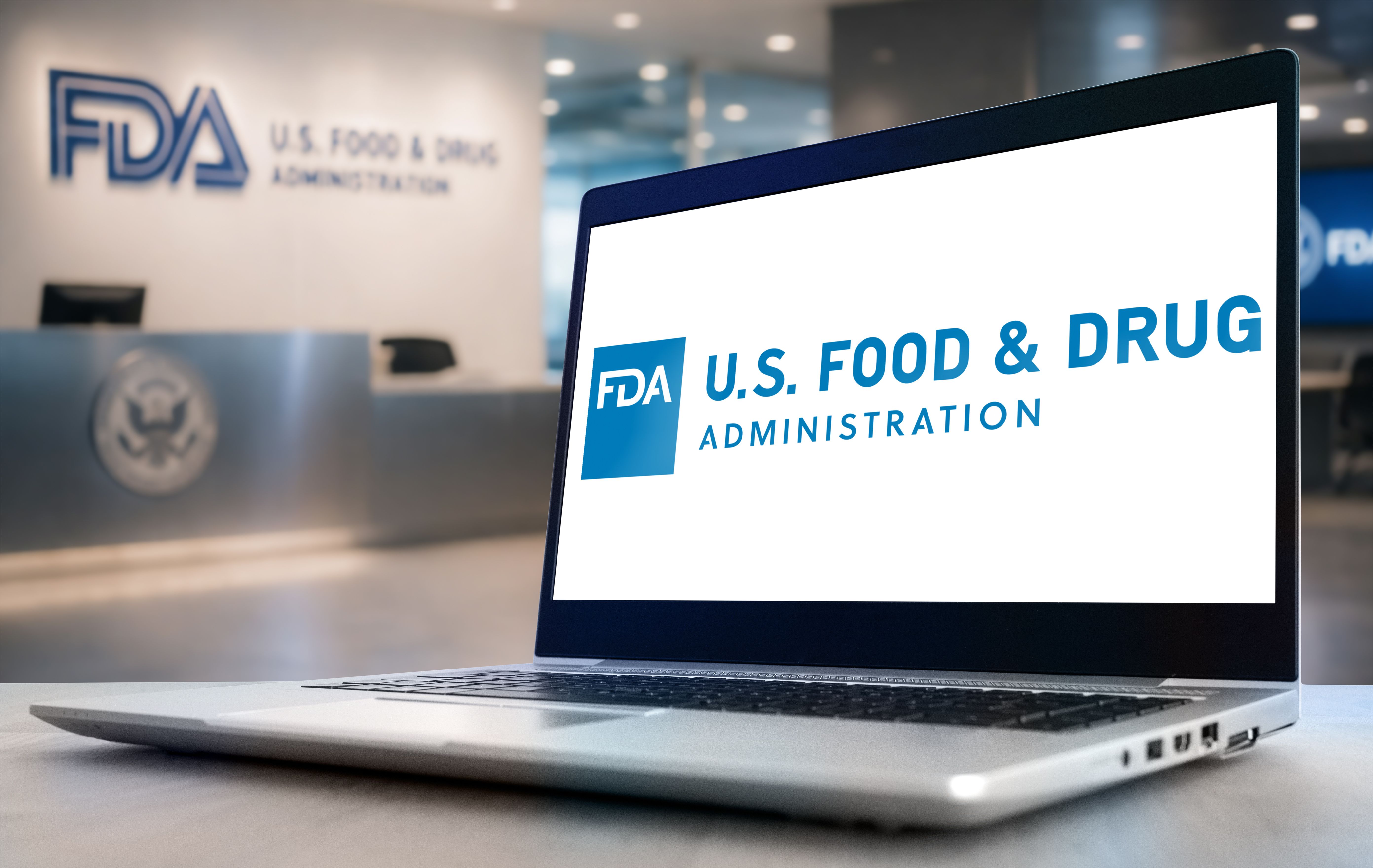 A laptop displaying the FDA logo, with FDA office signage visible in the background. | Image Credit: © gguy - stock.adobe.com