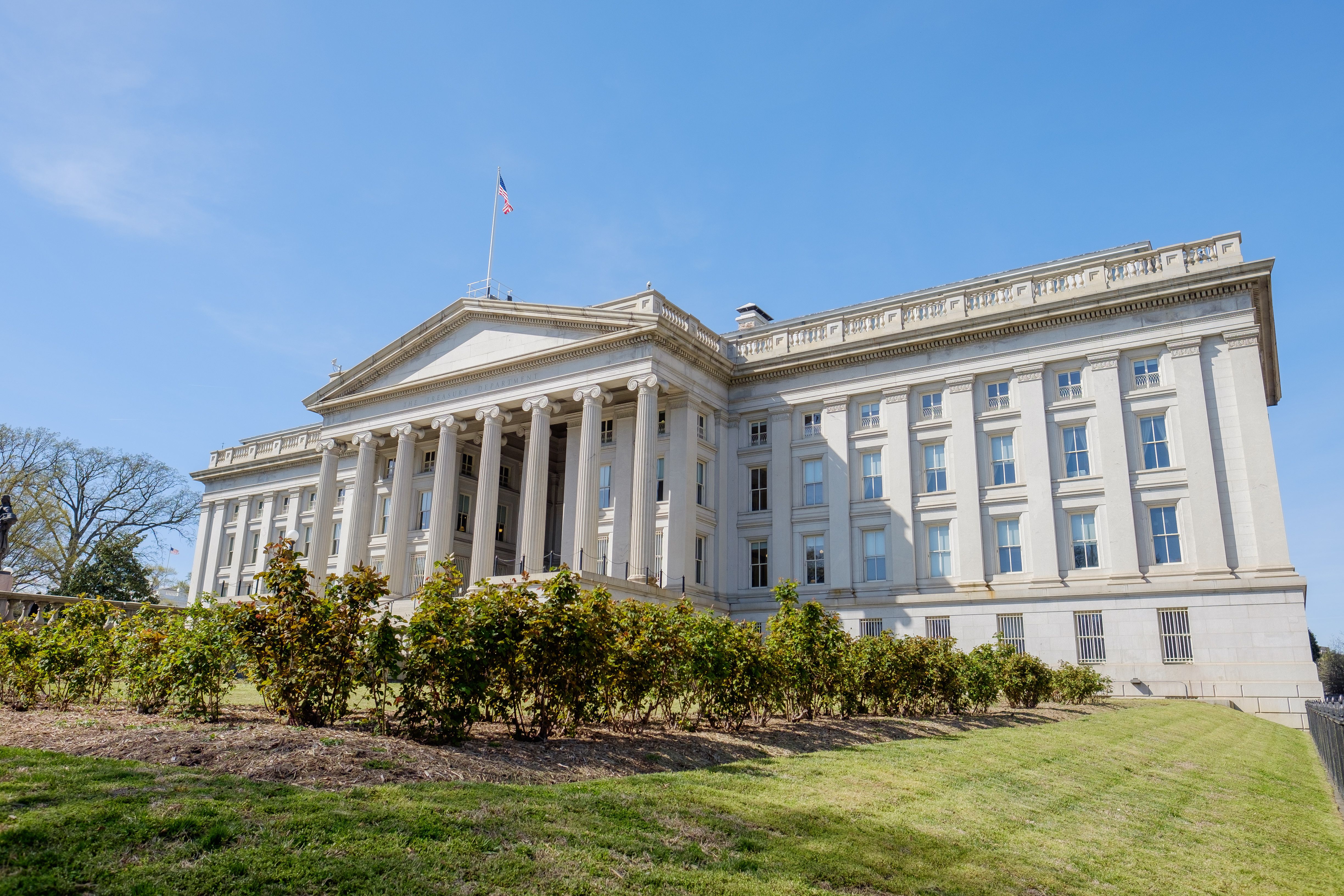Exterior of the US Department of the Treasury building in Washington, DC, with a columned entrance, an American flag on the roof, green shrubs and a lawn in the foreground, and a clear blue sky. | Image Credit: © Jorge Moro - stock.adobe.com
