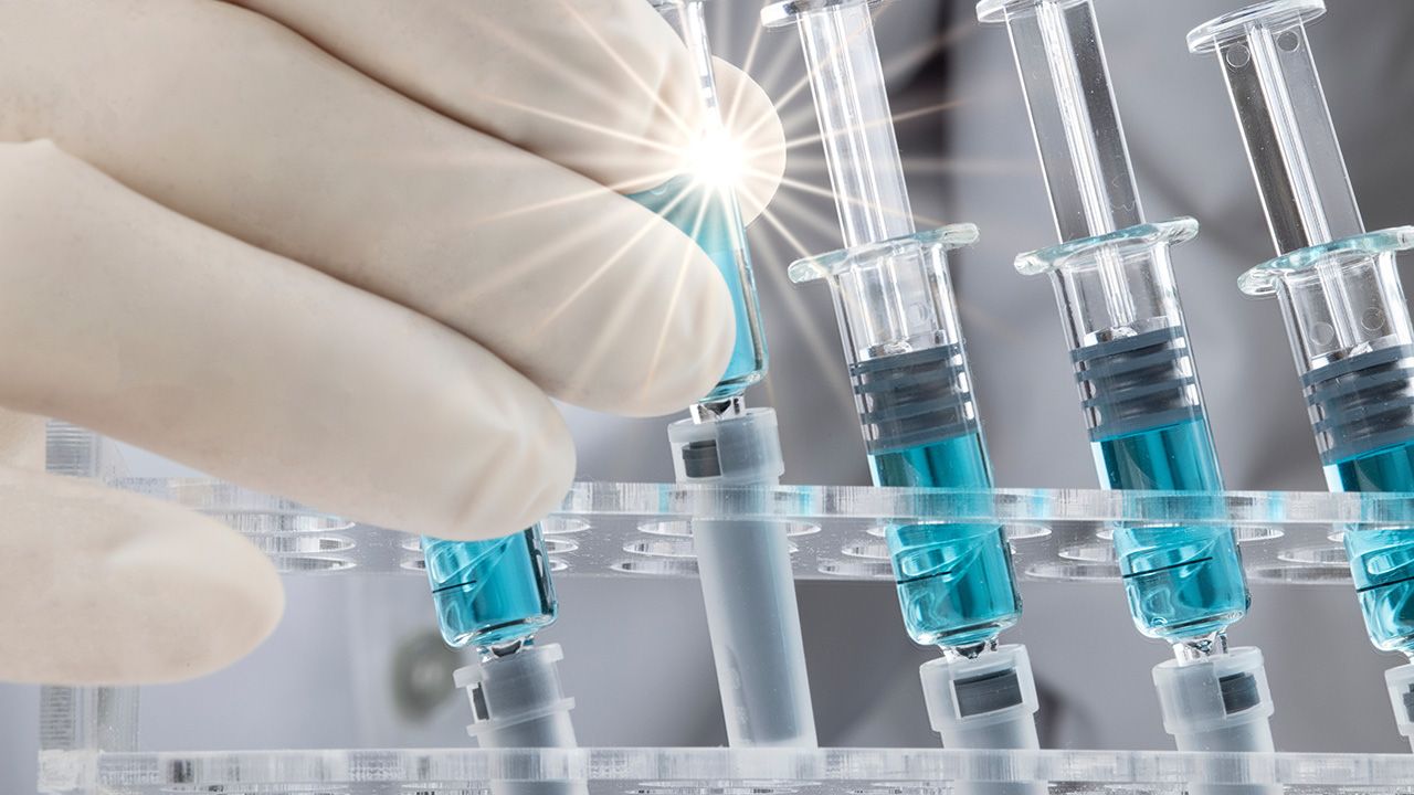 Closed up image of a doctor holding a glass syringe with several other syringes prefilled with blue liquid on a rack. Covid-19 vaccination concepts. | Image credit: © SOPONE - © SOPONE - stock.adobe.com 